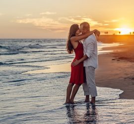 Un couple enlassé sur le bord d'une plage. Elle porte une robe rouge et lui est habillé de blanc.